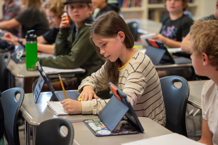 A student in a striped sweater writes in a notebook while using a tablet in a classroom filled with peers.