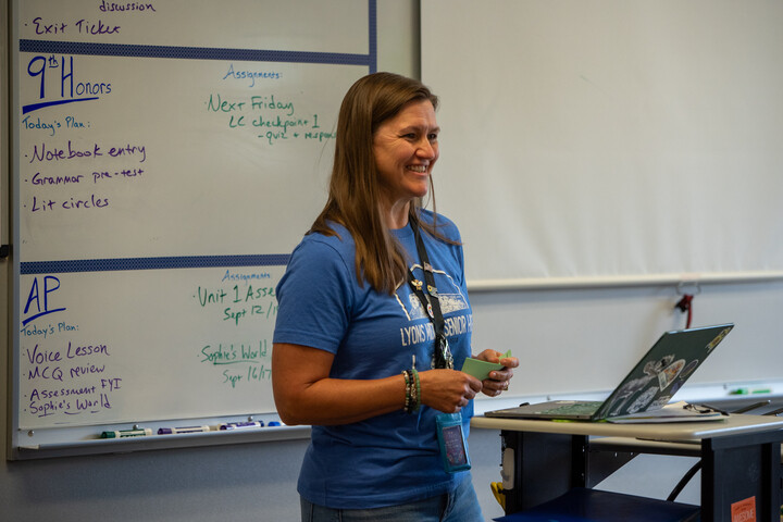 A smiling teacher stands in front of a classroom whiteboard, discussing plans with students while holding a card.