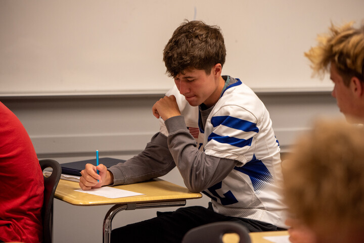 A student in a football jersey sits at a desk, writing on paper with a focused expression in a classroom setting.