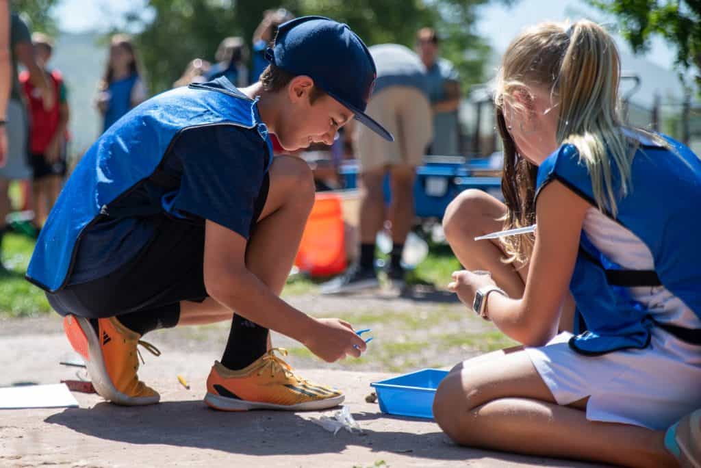 Male elementary student squatting down on left with two female students sitting on the ground on the right examining water samples. 